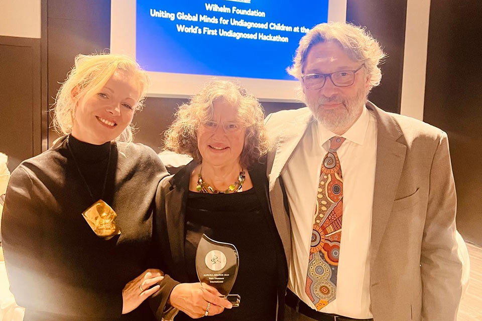 Alexandra Heumber Perry stands with Helene Cederroth, holding the glass Aurora Award, and Mikk Cederroth in front of a Wilhelm Foundation presentation slide.