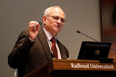 Bill Gahl, direktör för NIH:s UDP i Bethesda, talar vid ett podium märkt Radboud Universiteit och håller upp ett litet föremål.