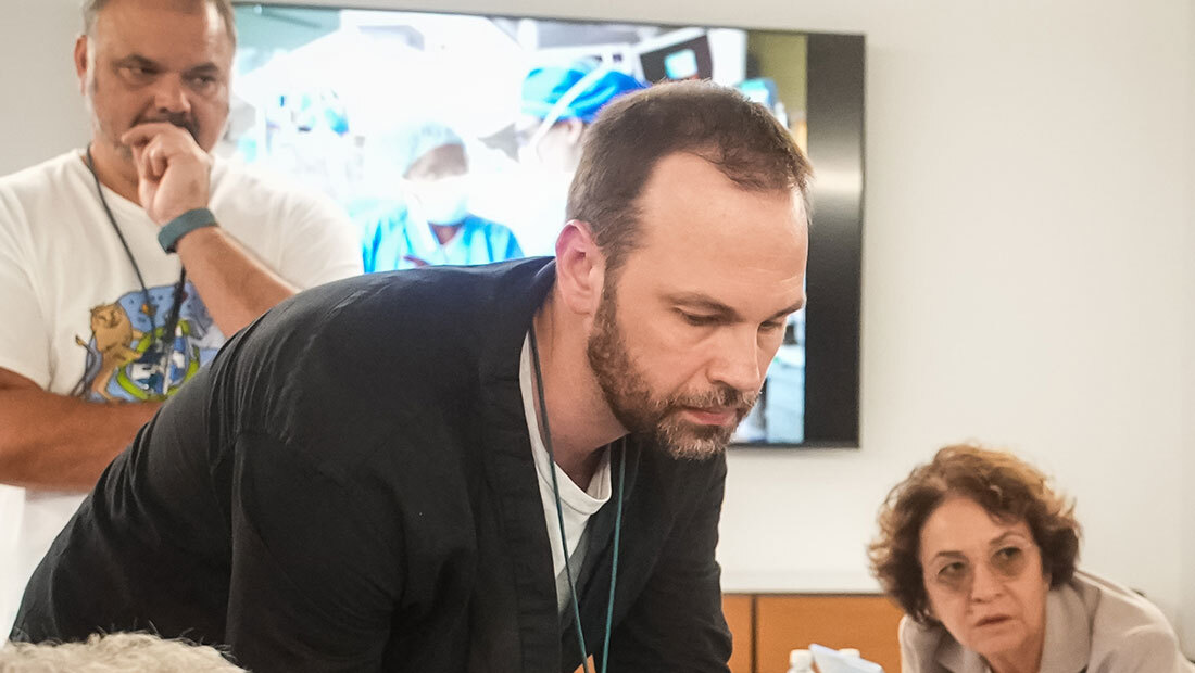 A clinician leans over a table in deep concentration during an Undiagnosed Hackathon, with colleagues watching an operating-room video feed behind him