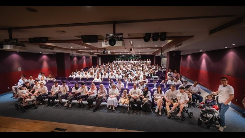 Group photo in an auditorium at the fifth Undiagnosed Hackathon in Singapore, September 2026: about 140 collaborators in white Undiagnosed Hackathon T-shirts fill the rows of purple seats, with the People Living With Undiagnosed Diseases and their families, several children in wheelchairs and strollers, in the front row.