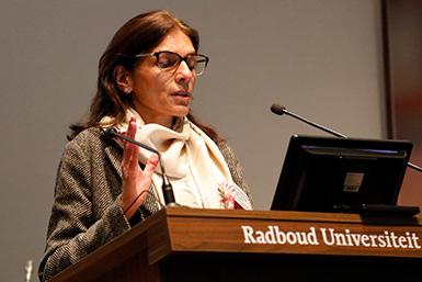 Professor Ratna Puri, i glasögon och vit scarf, talar vid ett podium märkt Radboud Universiteit och gestikulerar med en penna.