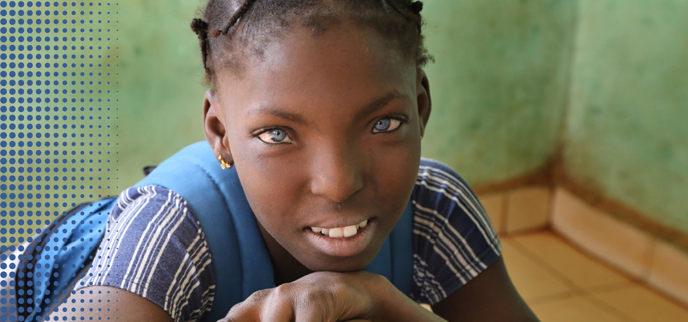 A smiling young girl with bright blue eyes rests her chin on her hands, framed by a blue dotted pattern.