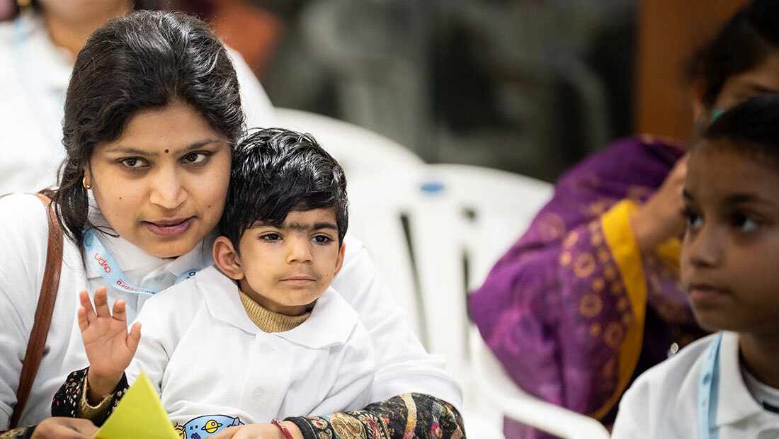 A mother holds her young son close during a gathering, both looking toward the front of the room