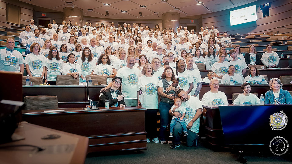 Large group photo of clinicians, researchers, data scientists, families, and collaborators gathered in a lecture hall, all wearing matching Undiagnosed Hackathon T-shirts. The image captures the full community of participants at the Undiagnosed Hackathon 2025, reflecting multidisciplinary collaboration, shared purpose, and collective commitment to solving complex undiagnosed diseases.