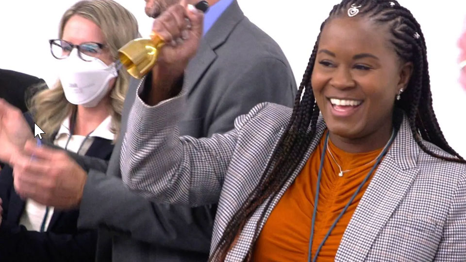 A smiling woman rings a small handbell during a group gathering, while people around her applaud. The moment captures celebration and shared joy, symbolizing a breakthrough or positive outcome being recognized collectively.
