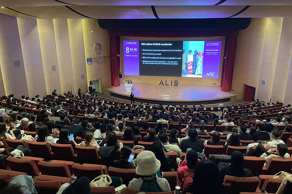 Wide view of a large auditorium filled with attendees seated in rows, listening to a speaker on stage presenting slides about people living with undiagnosed diseases. A large screen displays statistics and images related to undiagnosed conditions, highlighting global impact during an international medical or scientific conference.