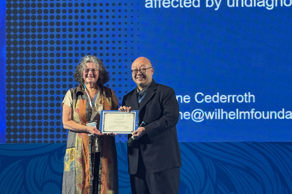 Two people stand on a conference stage holding a framed certificate and smiling toward the camera. They are positioned in front of a large blue presentation screen with text related to undiagnosed diseases, capturing a moment of recognition during an international genetics or medical conference.