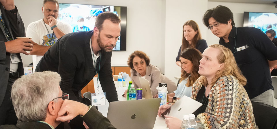 A diverse group of clinicians, researchers, and data experts gathered around a table during an Undiagnosed Hackathon, collaboratively reviewing a complex case on laptops and tablets, engaged in focused discussion and problem-solving to advance diagnosis for an individual with an undiagnosed disease.
